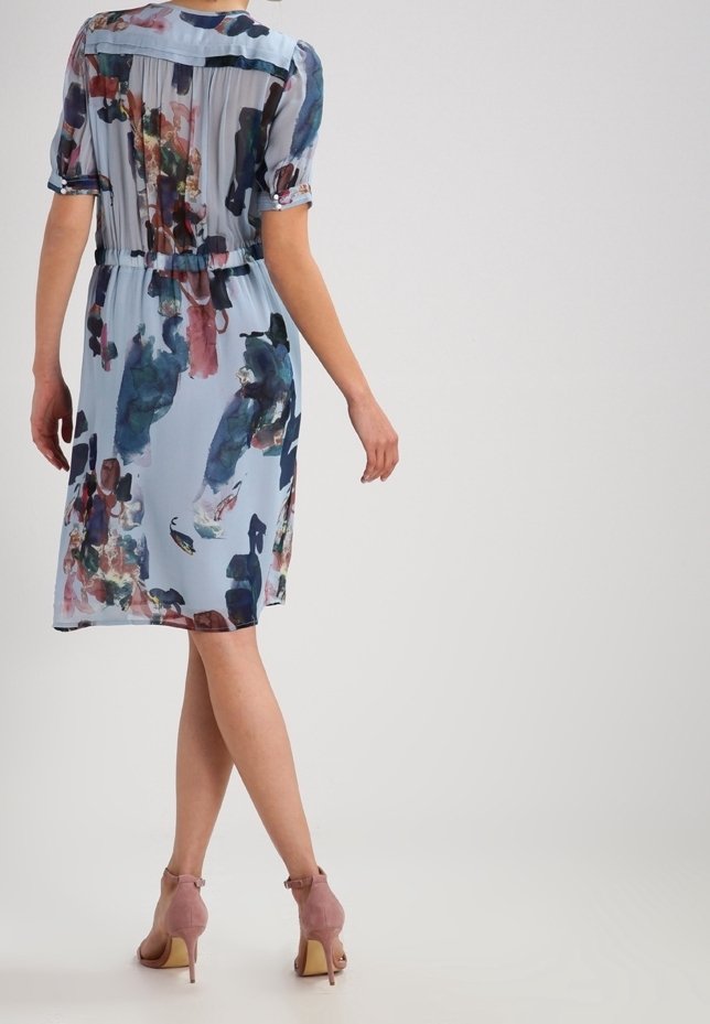 Femme portant une robe fleurie bleu clair longueur genoux avec manches courtes et des chaussures roses à talons hauts à bride cheville, debout sur un fond uni.