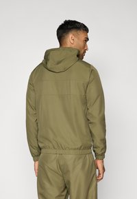 Veste à capuche vert olive avec des poignets et une taille élastiques, texture lisse et un design simple. Présente un détail de couture subtil dans le dos.