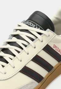 Acercamiento a una zapatilla Adidas Spezial crema con rayas negras, costuras rojas, cordones blancos, etiqueta negra en la lengüeta y suela de goma.