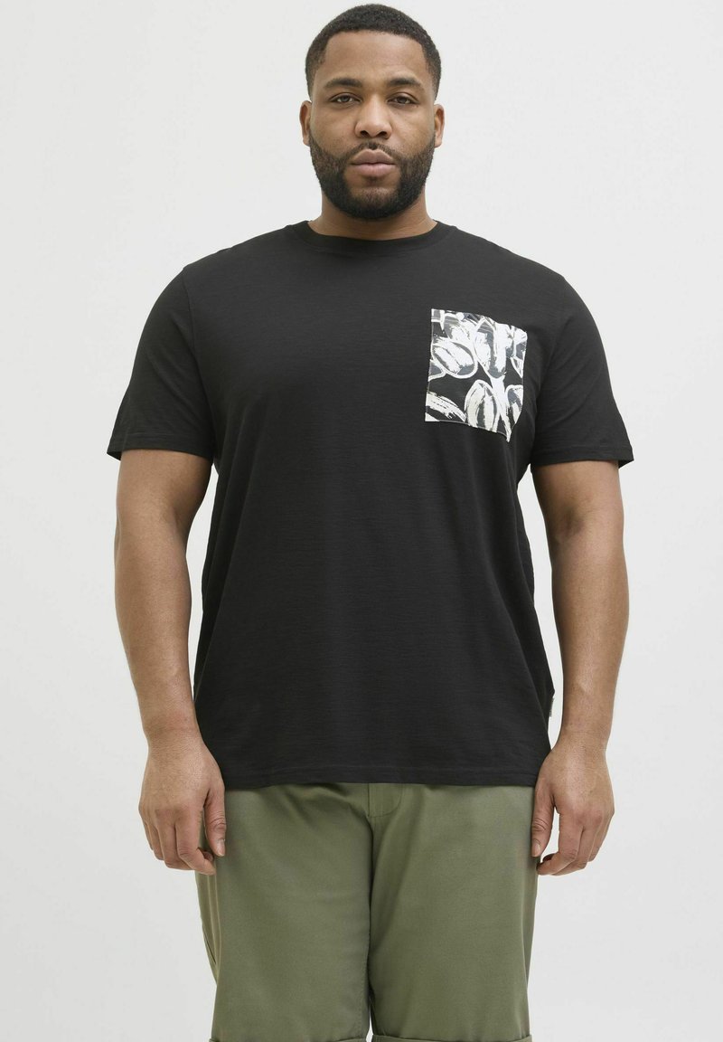 T-shirt noir à manches courtes, avec une poche à motif blanc. Le matériau semble être du coton ; porté avec un short vert olive.