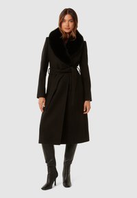 Abrigo de lana negro con un gran cuello de piel sintética, cintura ceñida, mangas largas y un silueta fluida. Llevado sobre botas negras hasta la rodilla.