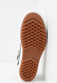 Gros plan sur la semelle marron à motif gaufre d’une chaussure Vans avec le logo Vans visible, chaussure posée debout contre une surface blanche.