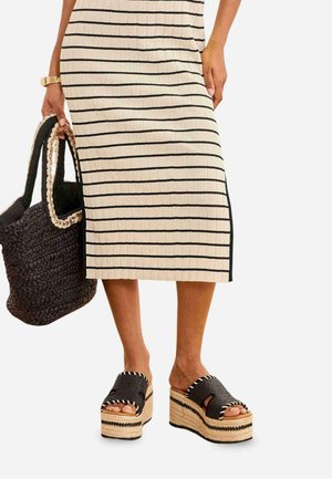 Mujer con vestido beige acanalado con rayas negras, sosteniendo un bolso negro tejido y llevando sandalias negras de plataforma con suelas trenzadas.