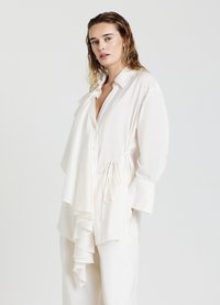 Donna con capelli biondi bagnati, che indossa una blusa bianca larga con un frontale arricciato e pantaloni bianchi abbinati, in piedi contro uno sfondo semplice.