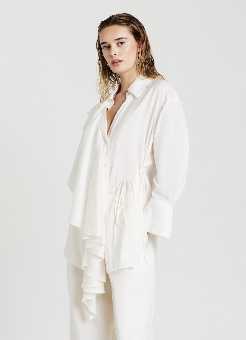 Donna con capelli biondi bagnati, che indossa una blusa bianca larga con un frontale arricciato e pantaloni bianchi abbinati, in piedi contro uno sfondo semplice.