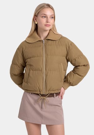 Jeune femme portant une doudoune beige avec un grand col et une jupe marron clair, debout, une main sur la hanche.
