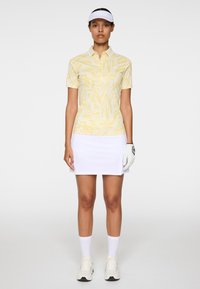 Golfowa koszulka polo w jasnym żółtym kolorze z abstrakcyjnym wzorem, zestawiona z białą spódnicą. Dodatki to biały daszek i rękawica golfowa.