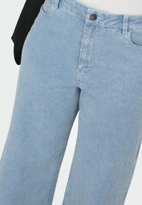 Gros plan sur un pantalon à rayures verticales bleu clair et blanc, porté avec un haut blanc rentré et une veste noire.
