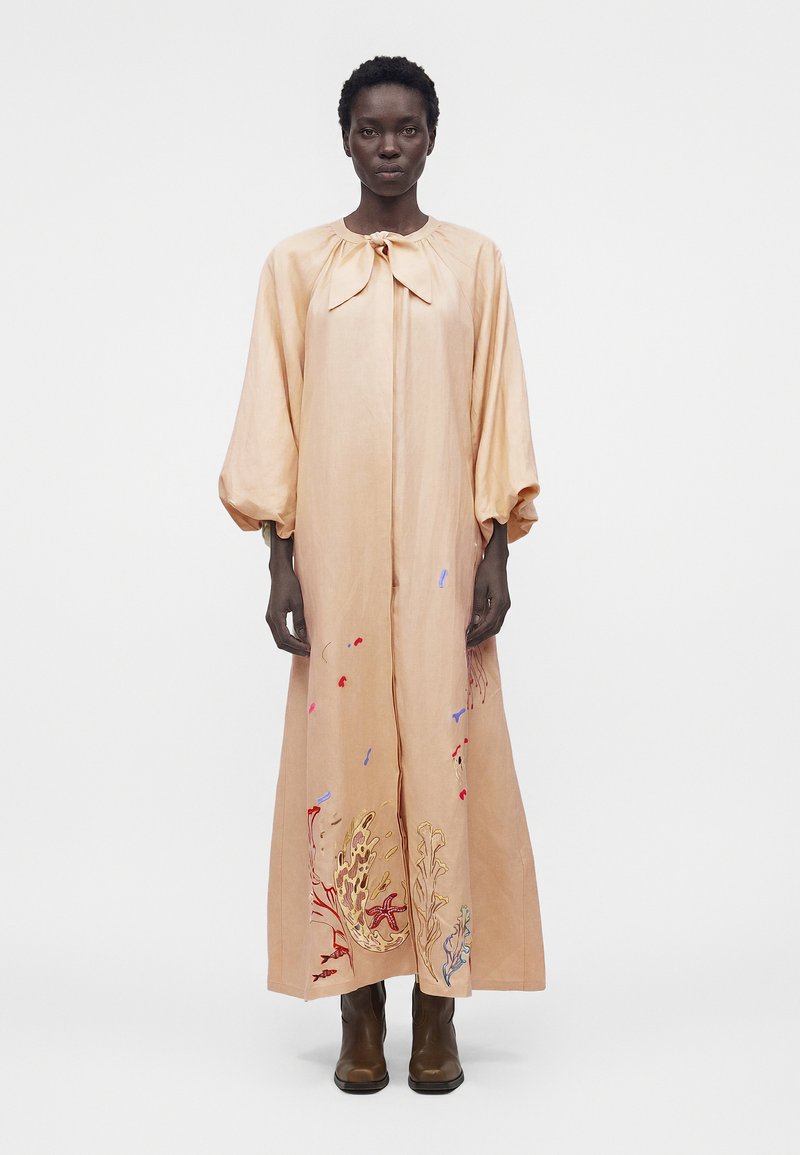 Γυναίκα που φοράει ένα μακρύ, ροδακινί φόρεμα με φουσκωτά μανίκια και κεντητά σχέδια κοραλλιογενών υφάλων κοντά στο τελείωμα, συνδυασμένο με καφέ μπότες.