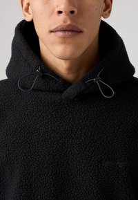 Sort fleece hættejakke med tekstureret overflade, der har justerbare snore og hætte, minimal branding synlig i matchende farve.