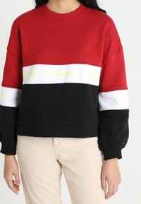 Felpa oversize rossa, nera e bianca con testo giallo "TOMMY". Presenta un collo a giro e maniche lunghe con polsini a coste.