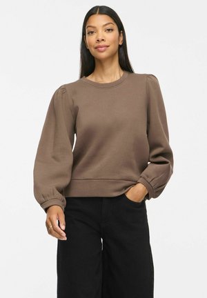 Kvinde med langt hår iført en brun langærmet top med puffede ærmer og sorte bukser, den ene hånd i lommen, neutral baggrund.