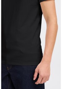 Tricou scurt cu mânecă neagră, dintr-un material neted, cu guler rotund, croială ajustată, purtat împreună cu blugi negri.