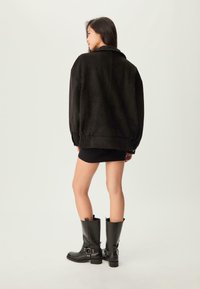 Femme aux longs cheveux noirs portant une veste en suède noire, une courte jupe noire et de hautes bottes en cuir noir, se tenant devant un fond uni.