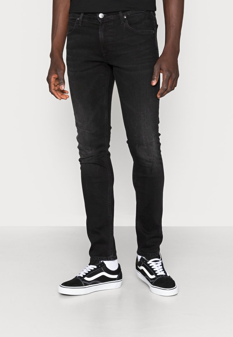 Homme portant un jean noir slim fit et des baskets noir et blanc, debout devant un fond blanc uni.