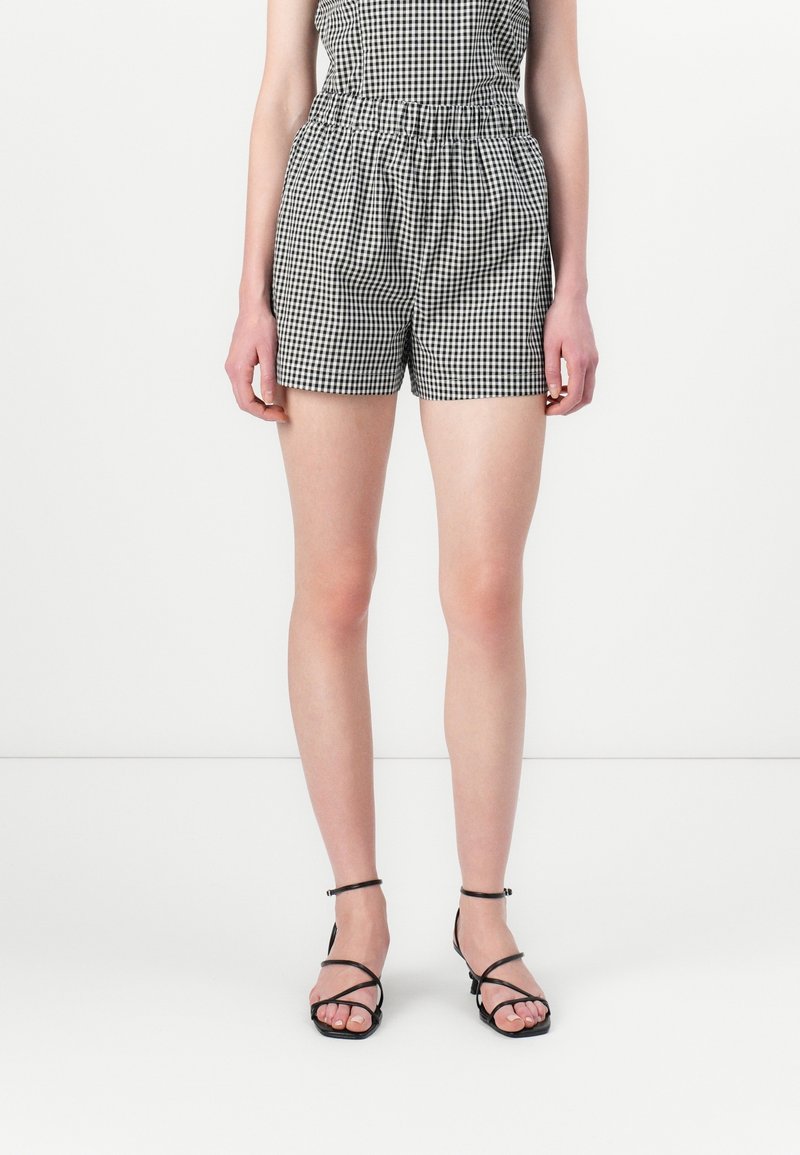 Femme portant une combinaison-short vichy noir et blanc et des sandales noires à talons et à brides, debout sur un fond blanc uni.