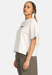 Jasno beżowy bawełniany t-shirt z krótkimi rękawami, okrągłym dekoltem, bocznymi rozcięciami i zielonym nadrukiem z przodu. Noszony z oliwkowymi spodniami.
