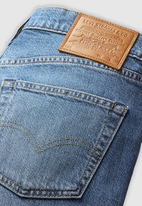 Denimjeans med en lys blå vask, med en lærrem på baksiden med preget merke, sømdetaljer og en buet lommeutforming.