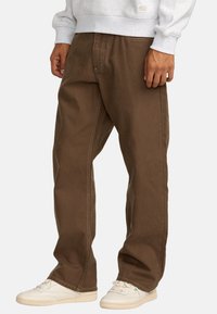 Donkerbruine, losse broek van katoen met contrasterende witte stiksels. Draagt een grijze sweatshirt en witte sneakers.