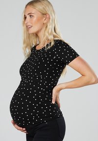 Schwarzes, kurzärmliges Schwangerschaftstop mit weißen Polka-Dots, das einen runden Ausschnitt und elastisches Material hat, das einen wachsenden Bauch accommodating.