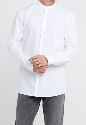Homme portant une chemise blanche à col mandarin avec des boutons cachés et un jean gris, ajustant la manche avec les mains.