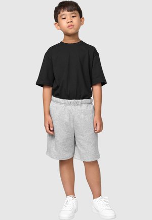 Jeune garçon debout portant un t-shirt noir oversize, un short gris avec taille élastique et des baskets blanches sur un fond clair uni.