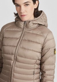 Vrouw die een beige gewatteerde pufferjack met capuchon en ritssluiting draagt, met een logo-opdruk op de bovenarm.