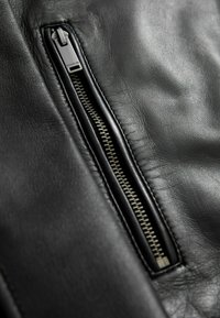 Detail einer schwarzen Lederjacke mit einem silbernen Reißverschluss auf der linken Seite. Die Oberfläche ist glatt, mit sichtbaren Nähten entlang der Kanten.