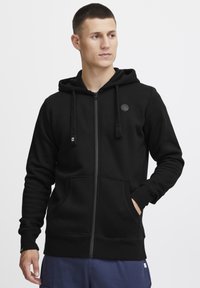 Haut à zip noir en tissu doux. Caractéristiques : capuche avec cordon de serrage, poches avant, poignets côtelés et logo circulaire sur la poitrine.