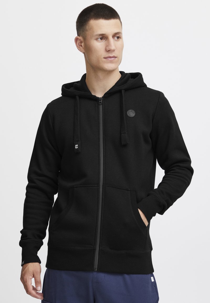 Haut à zip noir en tissu doux. Caractéristiques : capuche avec cordon de serrage, poches avant, poignets côtelés et logo circulaire sur la poitrine.