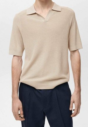 Mężczyzna w beżowej, krótkorękawowej, dzianinowej koszulce polo wpuszczonej do ciemnogranatowych spodni, stojący ze swobodnie opadającymi ramionami.