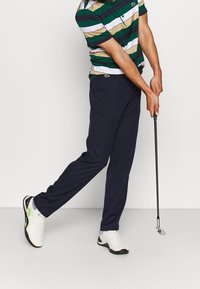 Marinblå golfbyxor och en flerfärgad randig polotröja. Tröjan har horisontella ränder i grönt, vitt och beige. Golfklubba i handen.