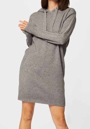 Kvinde iført en grå strikket hættetrøjekjole med ribbet manchetter og kant, hænderne foldet, stående mod en ensfarvet lys baggrund.