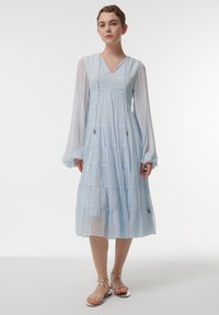 Robe midi bleu clair à volants avec de longues manches transparentes et un décolleté en V. Présente des embellissements subtils sur le corsage. Associée à des sandales métalliques.