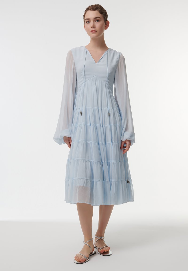 Robe midi bleu clair à volants avec de longues manches transparentes et un décolleté en V. Présente des embellissements subtils sur le corsage. Associée à des sandales métalliques.