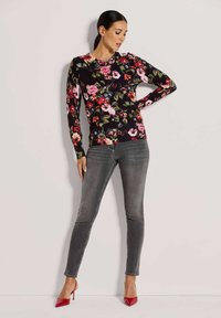 Strickjacke mit floralen Mustern in schwarzem Strick, mit langen Ärmeln und rundem Ausschnitt. Kombiniert mit grauen Skinny-Jeans und roten High Heels.