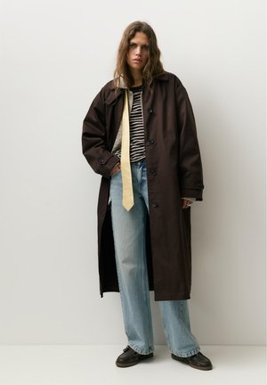 Personne portant un long manteau sombre, une chemise rayée, une cravate jaune, un jean bleu clair et des chaussures noires, debout devant un fond clair uni.