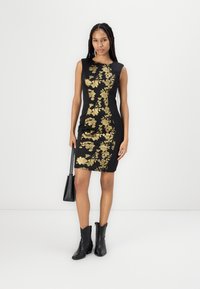 Rochie neagră ajustată cu broderie florală aurie, design fără mâneci. Modelul poartă cizme negre până la gleznă și ține o posetă mică neagră.