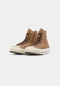Brune høye sneakers laget av lær med en kremfarget gummisåle, med brune snøringer og en rund logo på siden.