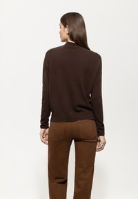 Frau mit langen braunen Haaren, die einen dunkelbraunen Pullover und passende braune Hose trägt, stehend mit dem Rücken zu einem schlichten weißen Hintergrund.