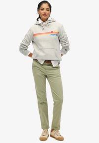 Felpa grigia con cappuccio, decorata da strisce colorate e logo, abbinata a pantaloni verde oliva. Il modello indossa sneakers beige con suole marroni.