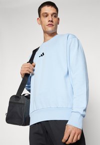 Lysblå sweatshirt med rund halsudskæring, ribbede manchetter og kant. Har et sort logo på brystet. Modellen holder en sort skuldertaske.