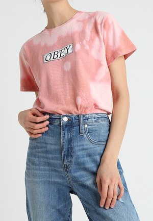 Roze tie-dye T-shirt met korte mouwen en een zwart "OBEY" logo op de borst, gecombineerd met een blauwe, hooggeremde spijkerbroek.
