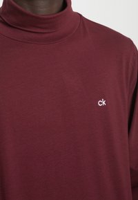 Burgundy polotröja i mjuk bomull, med en subtil vit "ck"-logotyp på vänster bröst. Slät textur, figurnära design.