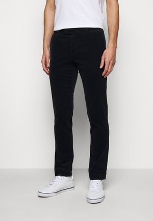 Mann trägt schmale schwarze Hose, weiße Turnschuhe und ein weißes T-Shirt, steht vor einem schlichten, hellgrauen Hintergrund.