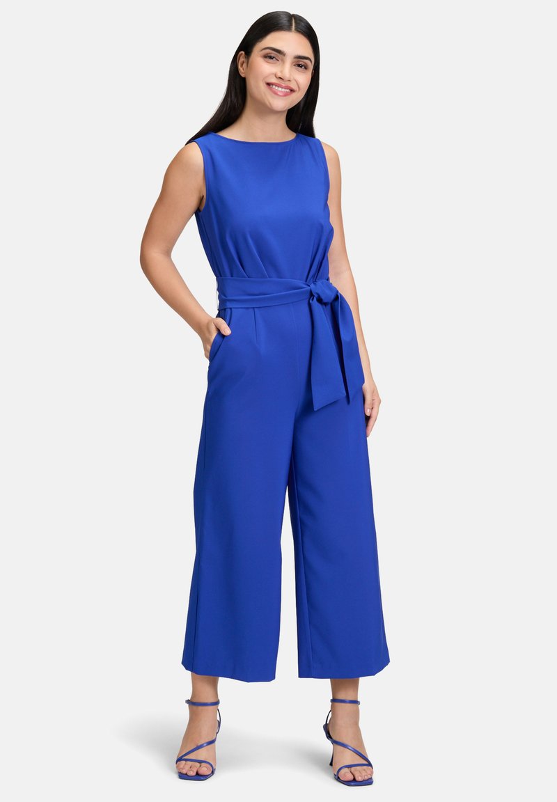 Modrá bezrukávová kombinéza s širokým strihom nohavíc, opaskom v páse a bočnými vreckami. Hladká látka s jednotnou farbou a minimálnymi akcentmi.