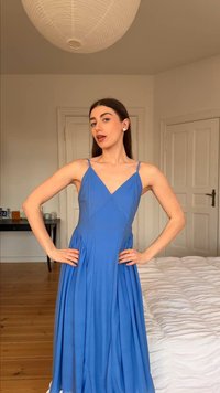 Γυναίκα με μακριά σκούρα μαλλιά που φοράει μπλε φόρεμα χωρίς μανίκια, στέκεται με τα χέρια στη μέση σε ένα υπνοδωμάτιο με ξύλινο πάτωμα και άσπρο πάπλωμα.