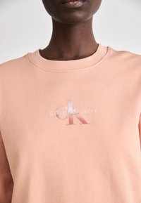 Sudadera de color melocotón hecha de una tela suave, con un cuello acanalado y un logotipo gráfico brillante que lleva el texto "Calvin Klein Jeans".