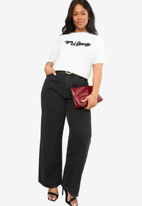 T-shirt blanc avec "Milano" en cursif noir, associé à un pantalon noir à taille haute et à larges jambes. Tenant une pochette bourgogne brillante et portant des talons noirs.