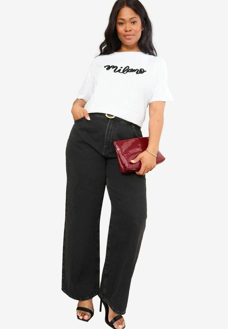 T-shirt blanc avec "Milano" en cursif noir, associé à un pantalon noir à taille haute et à larges jambes. Tenant une pochette bourgogne brillante et portant des talons noirs.